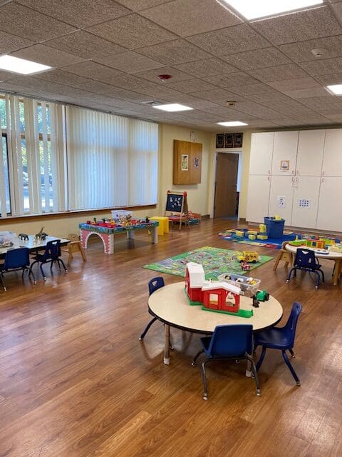 Children's playroom with tables, chairs, and toys, including a train set and building blocks.