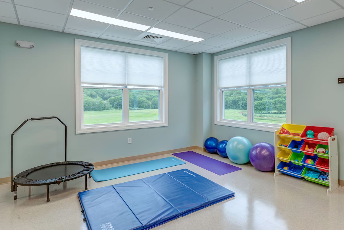 Room with therapy and exercise equipment including mats, balls, a trampoline, and shelves of toys.
