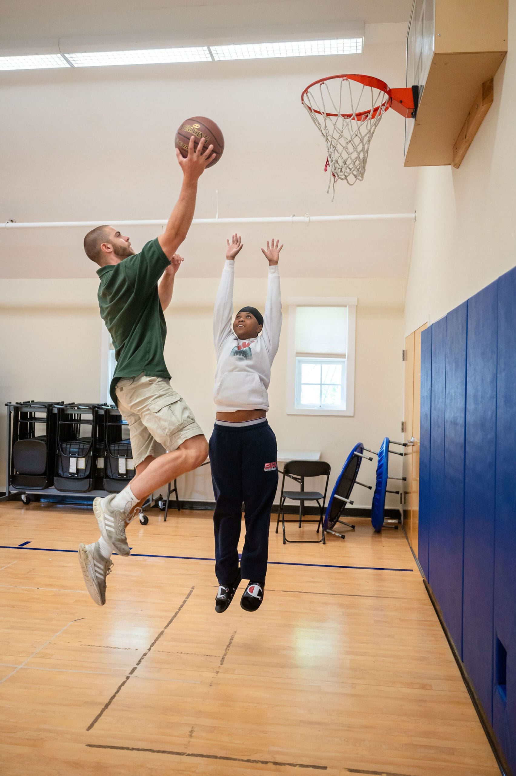 Man and girl playing basketball indoors, man jumping with a ball, girl reaching for the hoop.