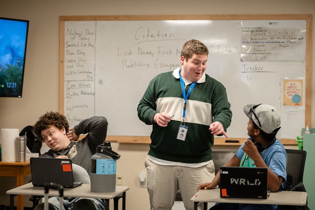 Person speaking to two children in a classroom with laptops and a whiteboard behind them.