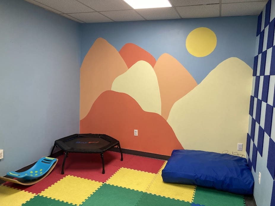 Room with colorful foam mat flooring, a trampoline, a blue bean bag, and a climbing sled.