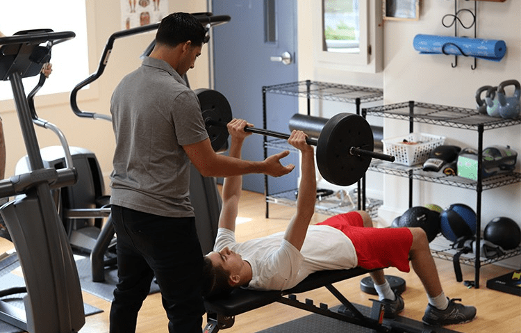 Person lying on a bench pressing a barbell while another person guides.