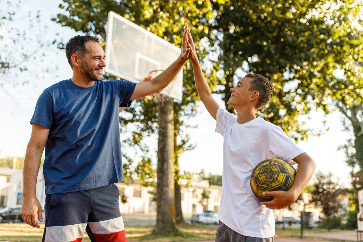 A man and a boy give each other a high five outdoors, with the boy holding a basketball.