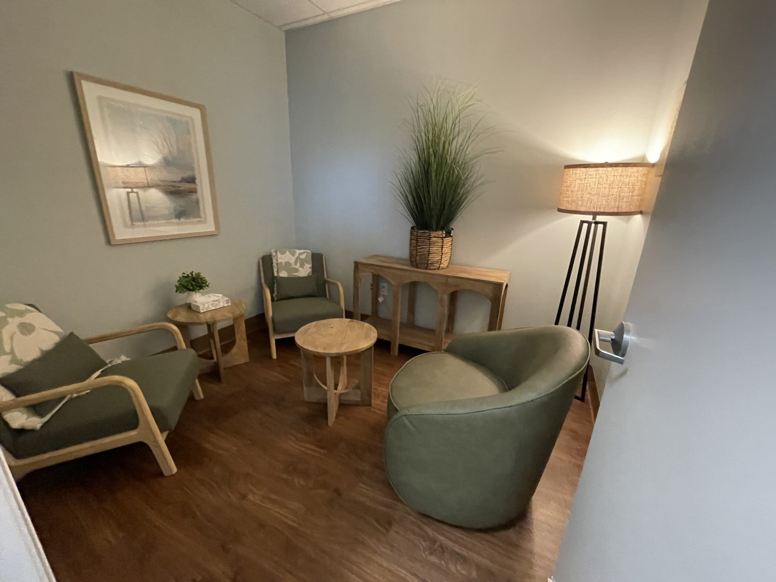 A cozy counseling room with green chairs, a wooden table, a large plant, and a floor lamp.