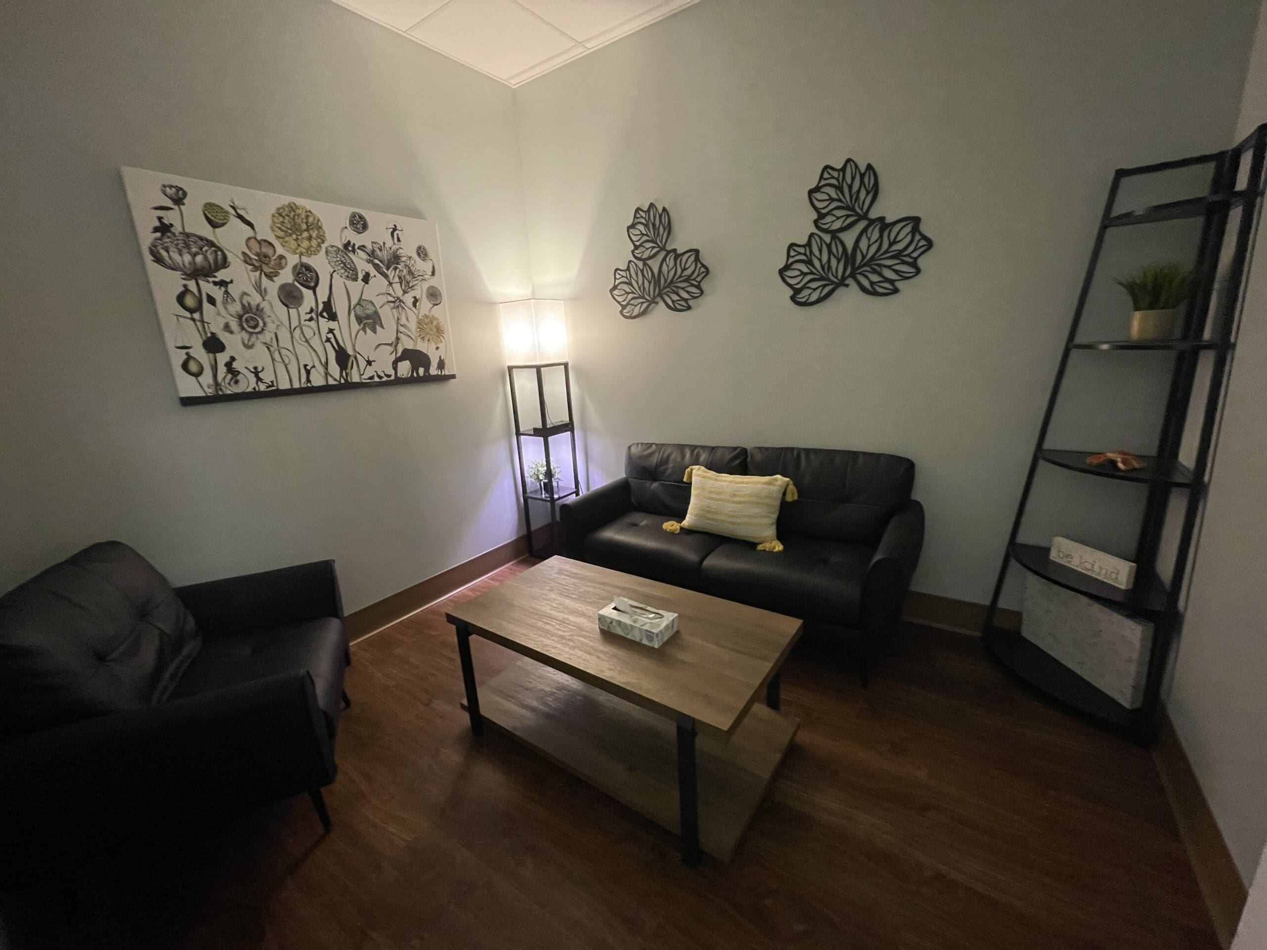 A cozy room with a black leather sofa, an armchair, a wooden coffee table, wall art, and a tall shelf with decor items.