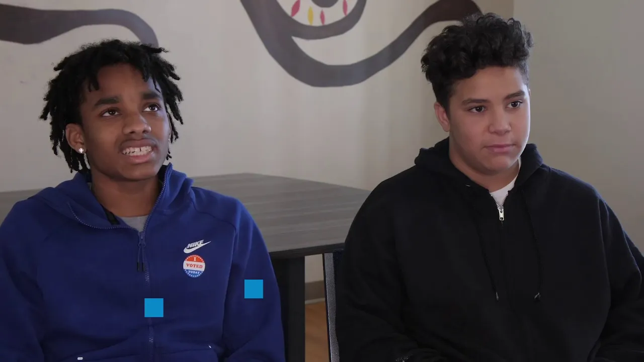 Two young people sit next to each other in a room, listening.