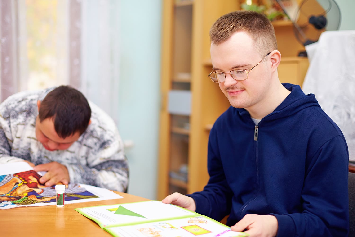 Student reading a book in a room with another one working on a project nearby.
