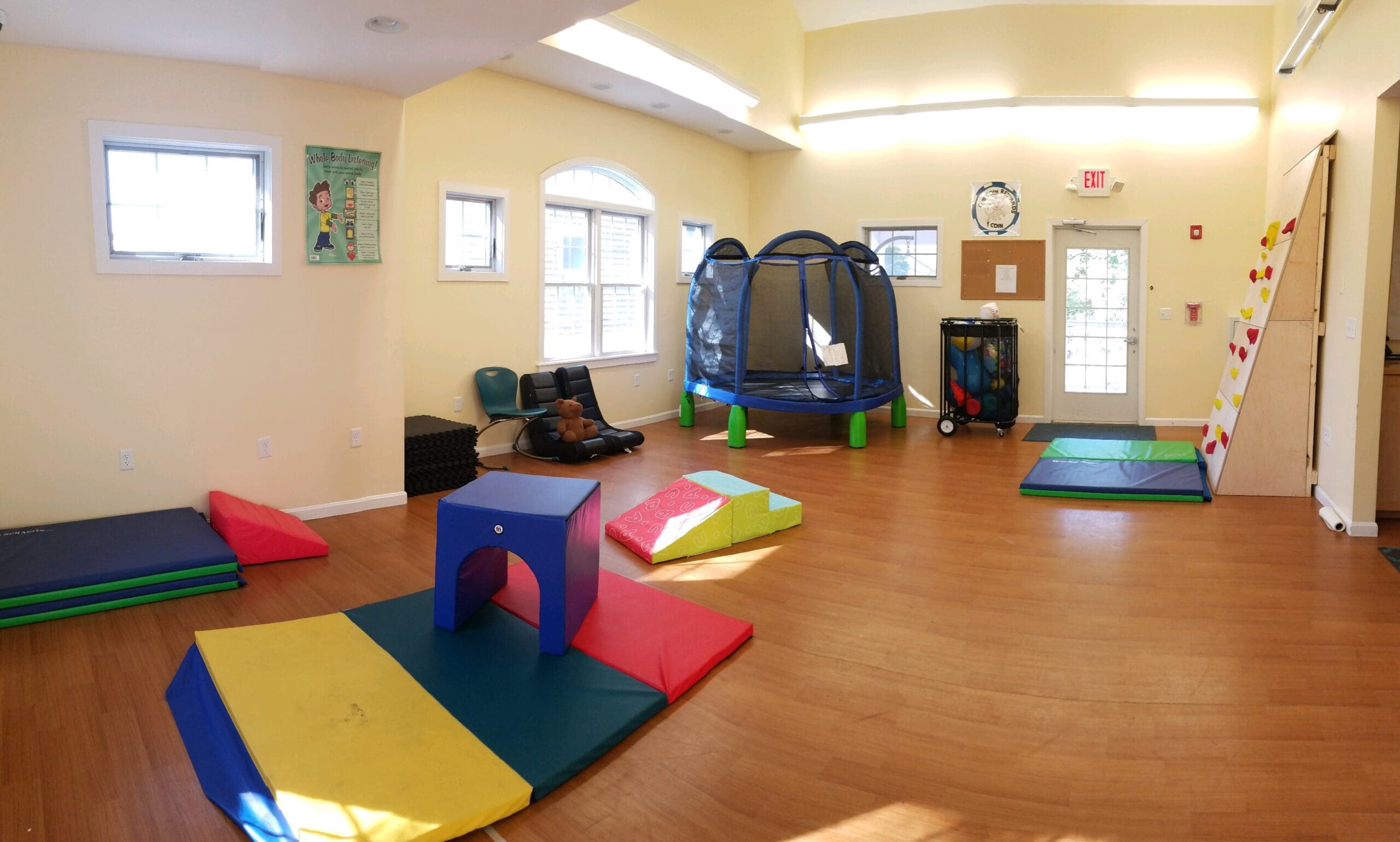 Indoor playroom with soft mats, small climbing wall, trampoline, and chairs.