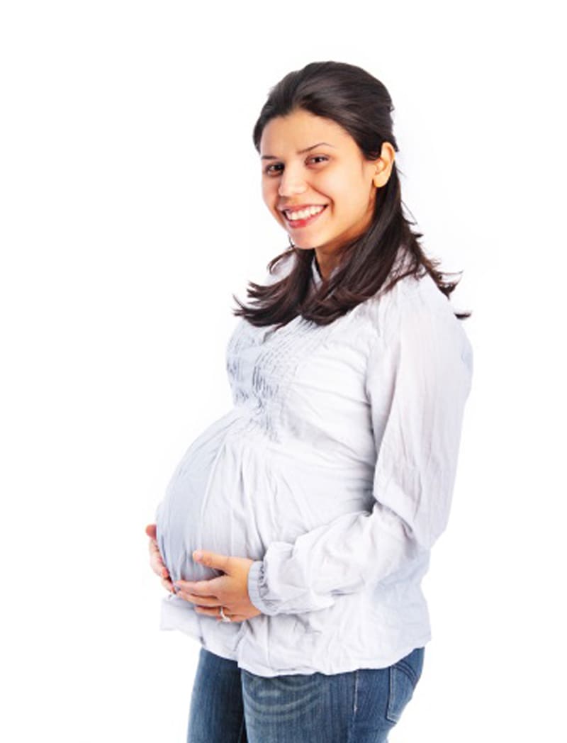 Woman with dark hair wearing a white shirt, holding her pregnant belly, smiling.
