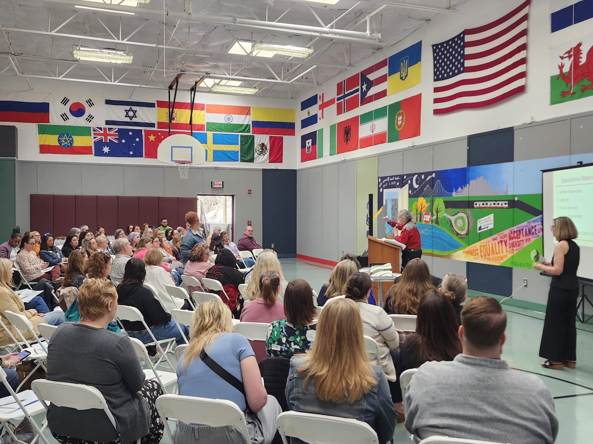 A large indoor gathering with many seated people listening to a speaker at a podium.