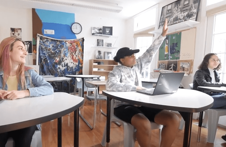 Students in a classroom, one raising hand, others seated at desks.