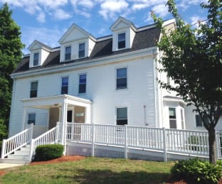 White multi-story house with a porch and surrounding trees.