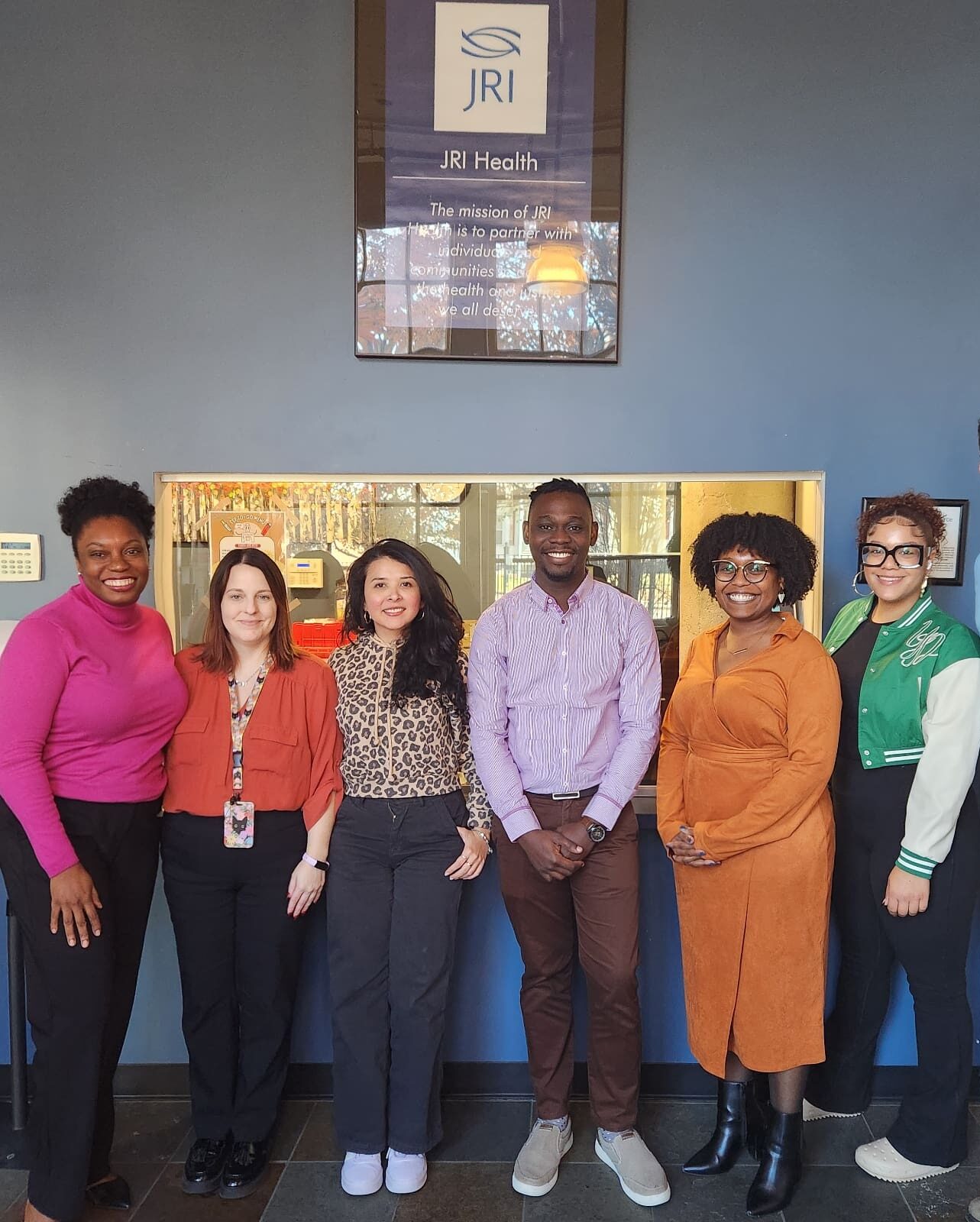 Group of Program Rise staff standing inside in front of a wall with a sign that says "JRI Health".