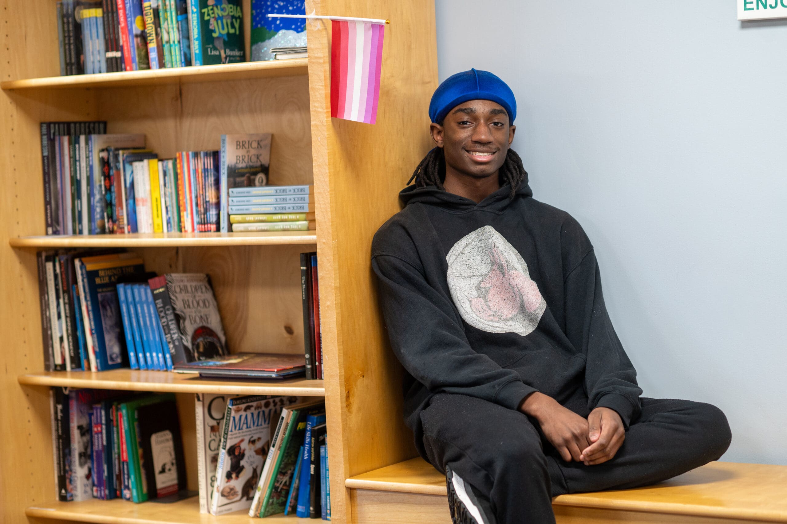 Young person sitting on a wooden bench with bookshelves nearby in a community space.