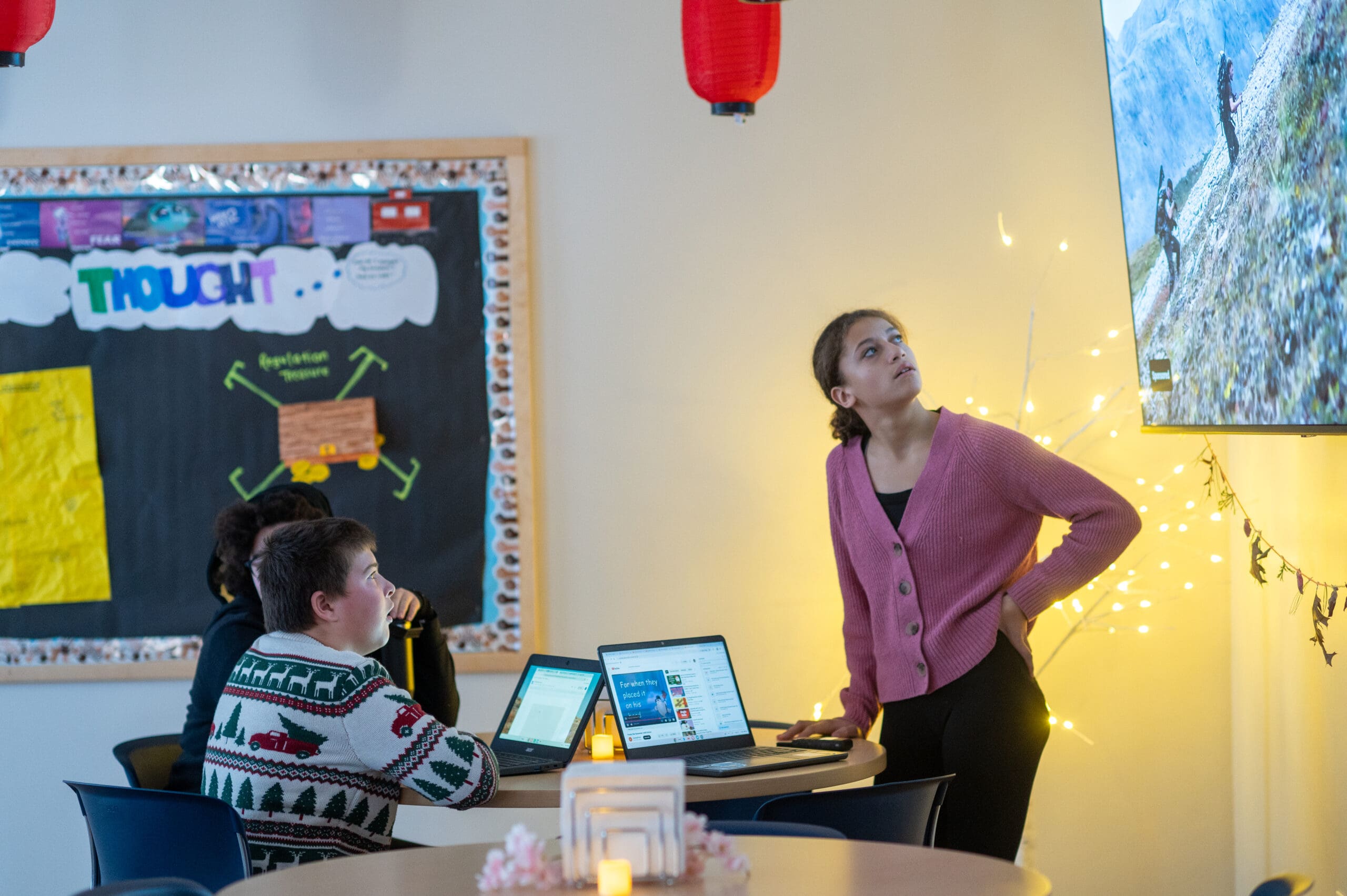 Teacher standing and talking to children watching a large screen in a classroom.