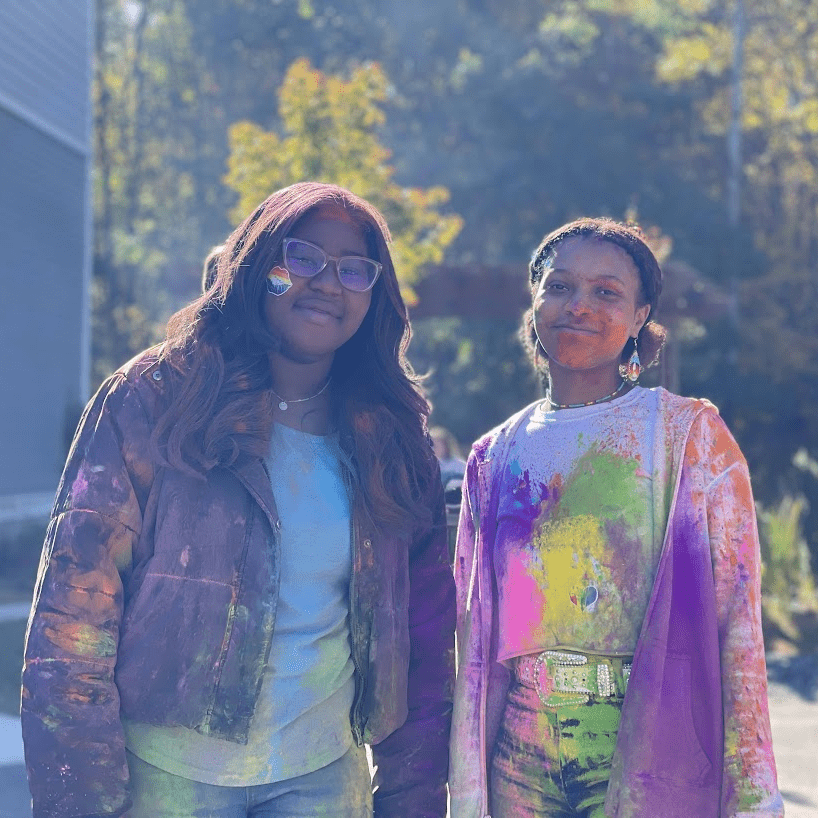 Two people with colorful clothing and paint on them, outdoors, smiling.