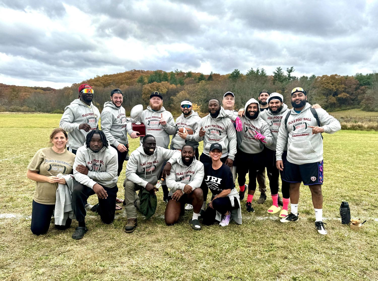 Group of people wearing "Flag Football" hoodies, some holding a trophy, outdoors.