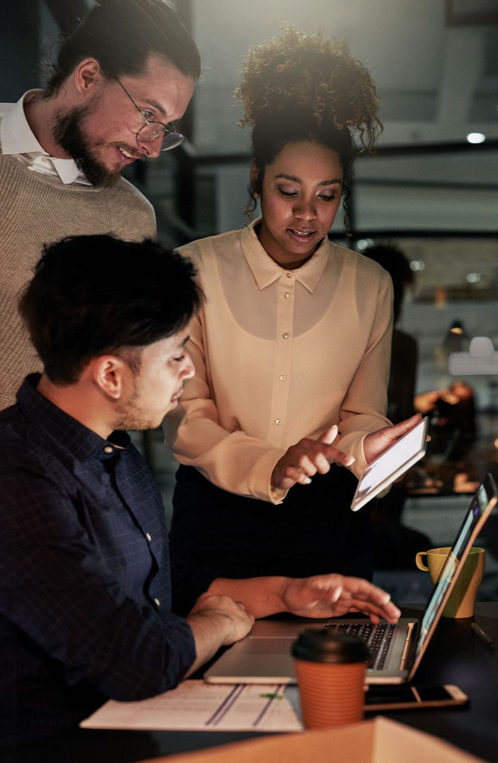 Three people gathered around a laptop and tablet in a dimly lit room.
