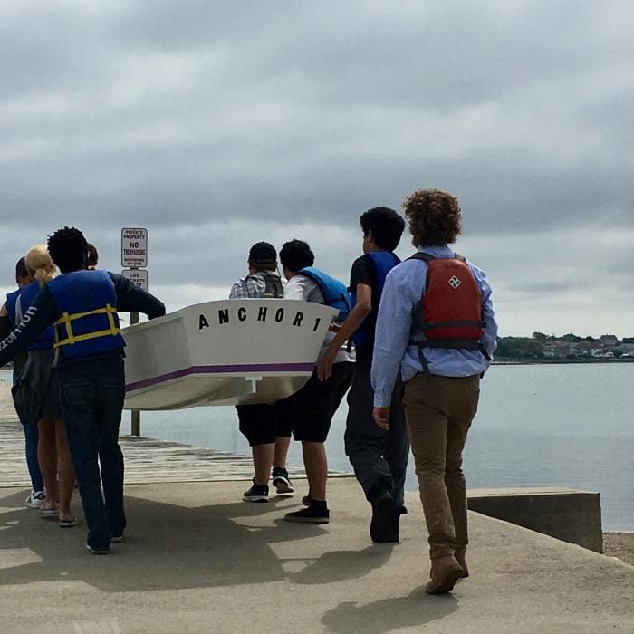 Group of people with life jackets carrying a boat named 'ANCHOR 1' by the water