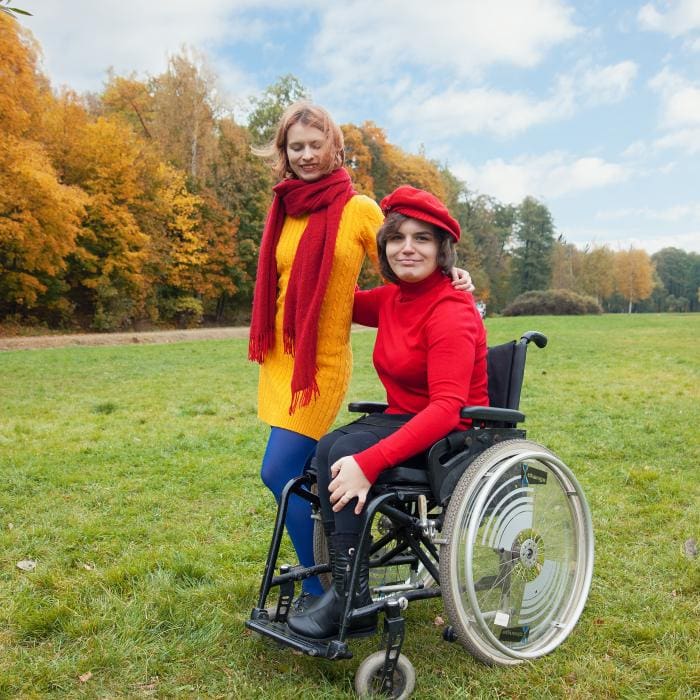 Two people outdoors, one in a wheelchair, both smiling, autumn trees.