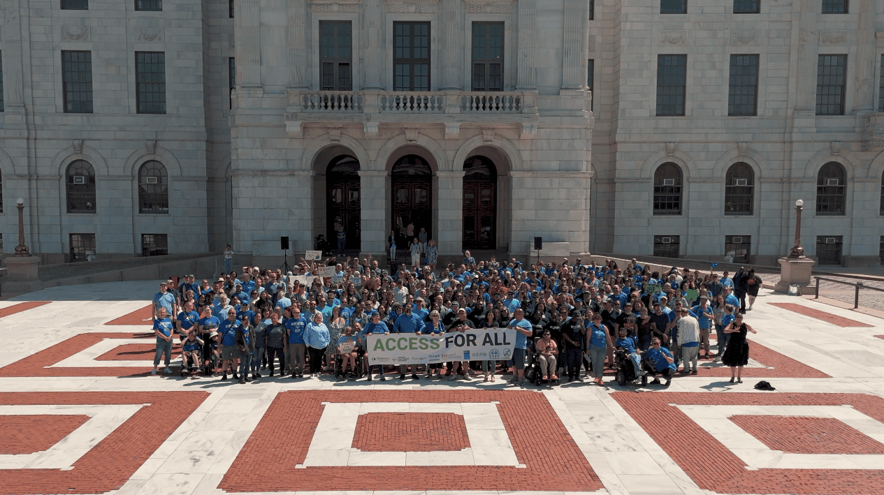 Large group of people holding a banner that reads "ACCESS FOR ALL" in front of a government building.