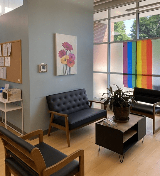 Waiting area with black and wooden chairs, potted plant, and rainbow flag.