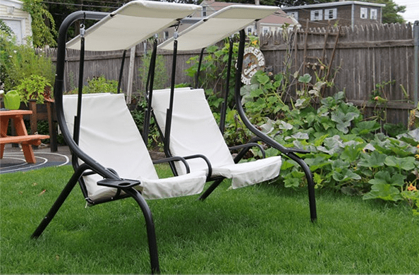 Two white outdoor swing chairs with canopy in a backyard garden with plants and a wooden fence