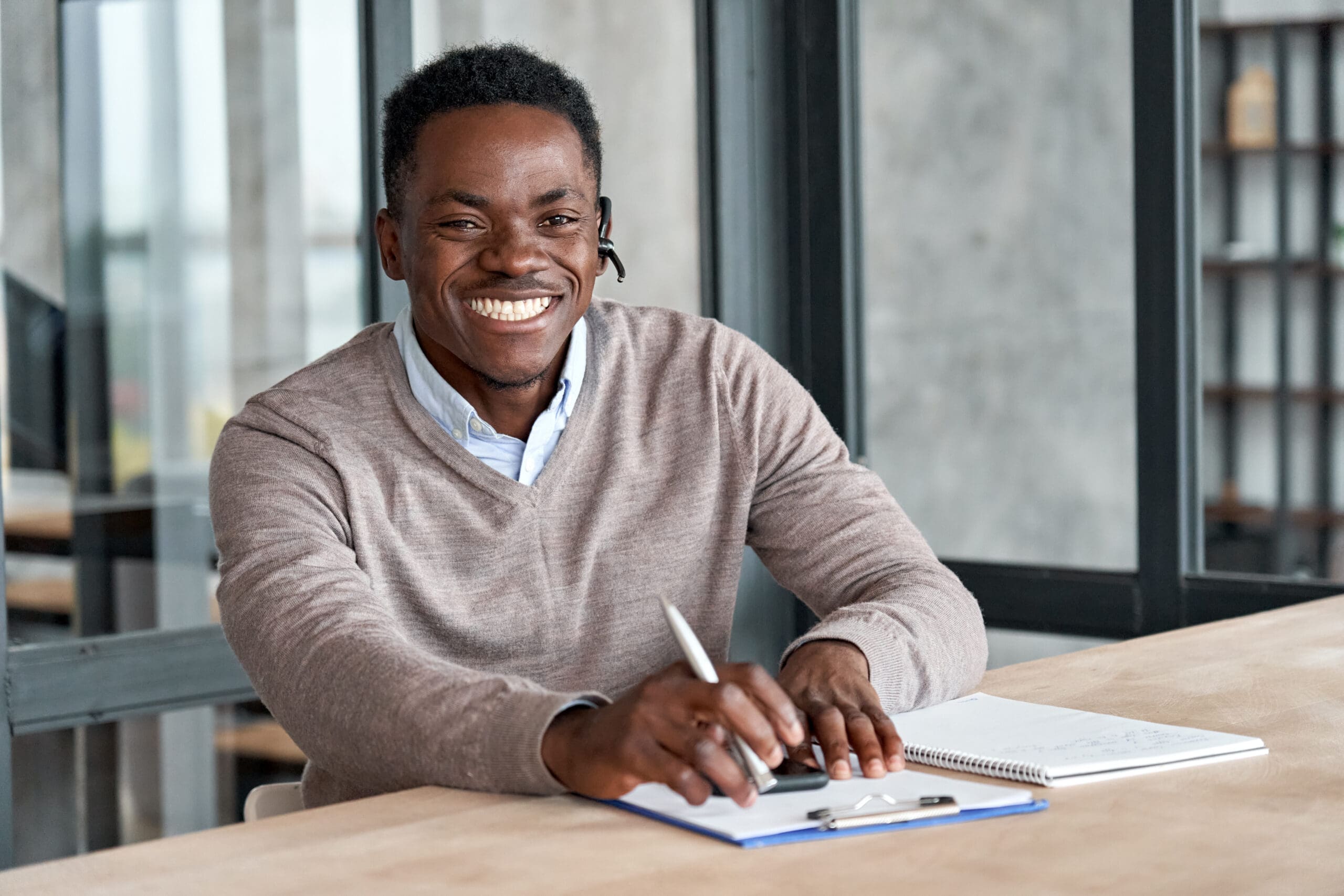 Man smiling, wearing a headset, sitting at a table with notebooks and a pen