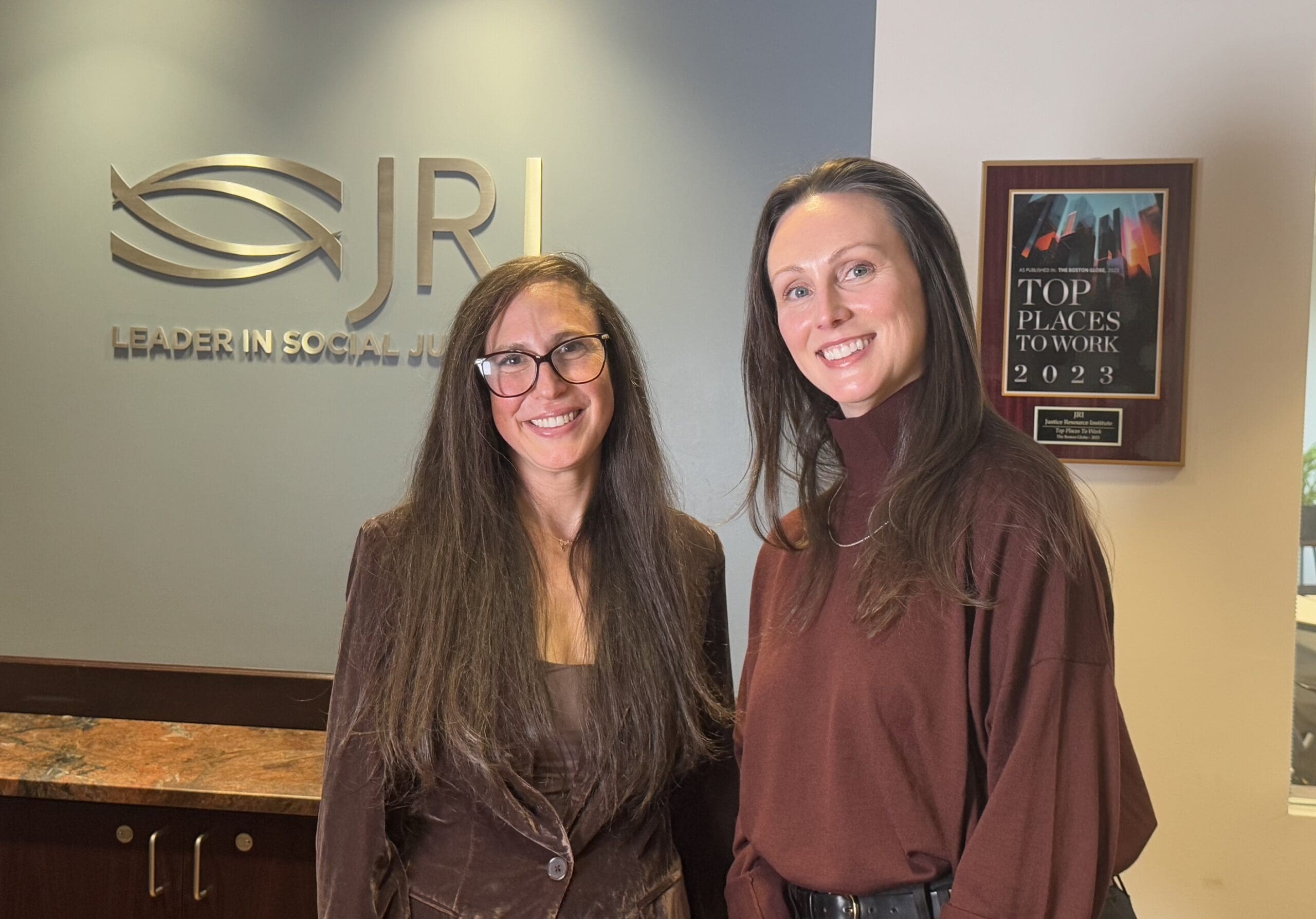 Two women smiling in front of a wall with a JRI logo and a plaque.