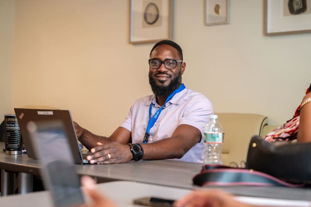 Man with glasses, beard, and light shirt sitting at a table with laptop.