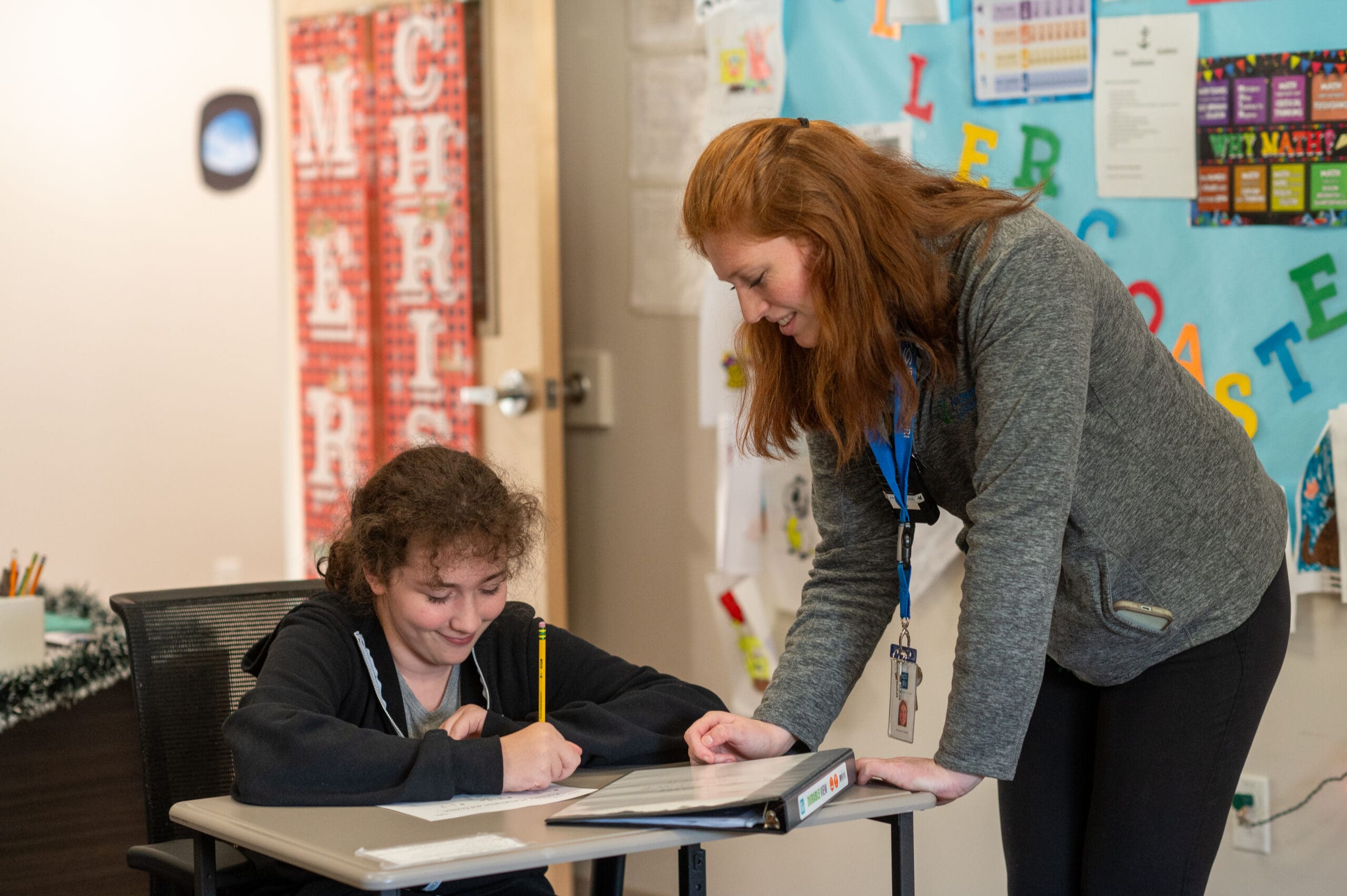A teenager sitting at a desk with a pencil, smiling as a woman leans over and points at her paper, in a classroom setting.