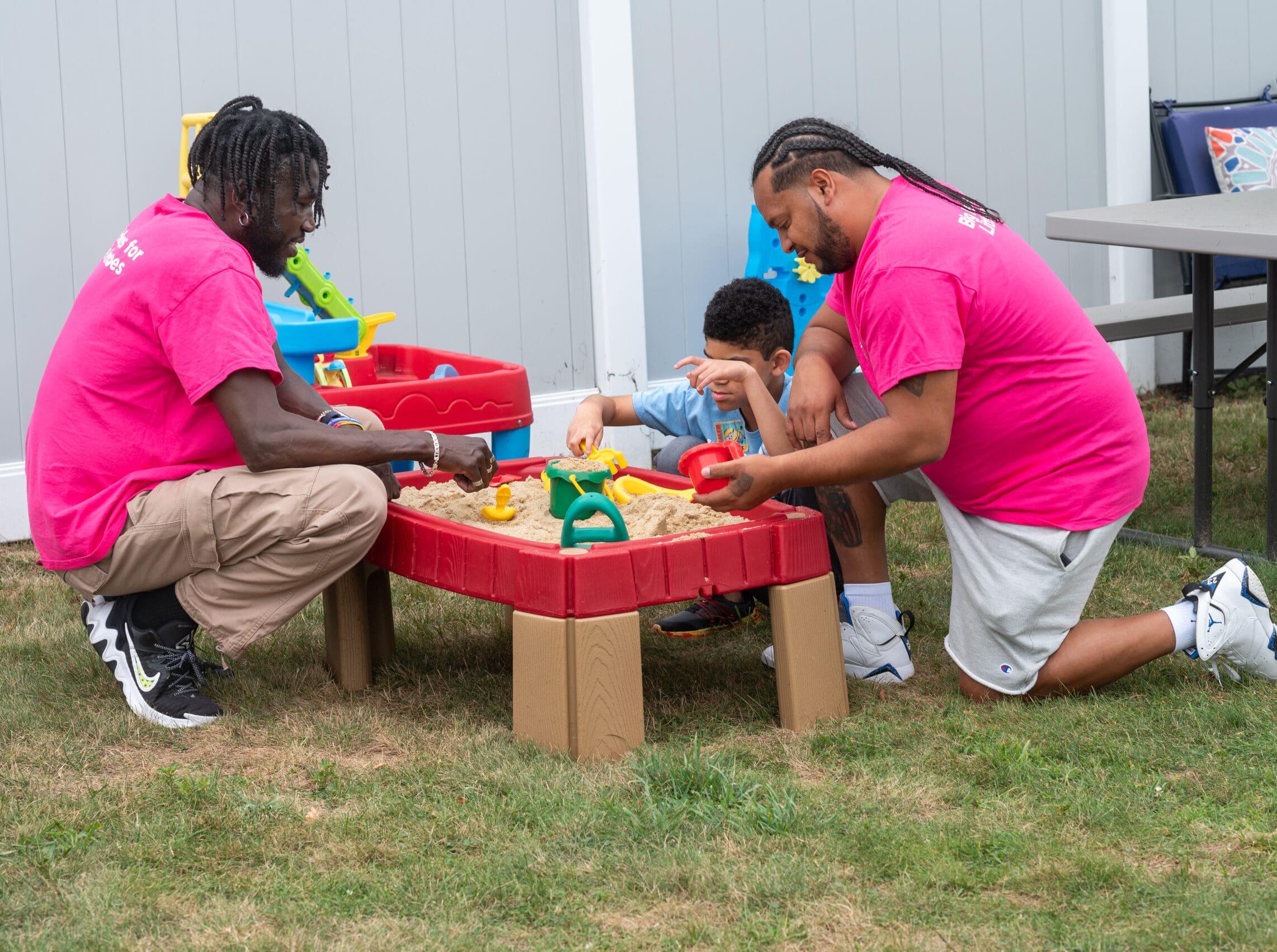 Two adults and a child playing in a sandbox outdoors with toy tools.