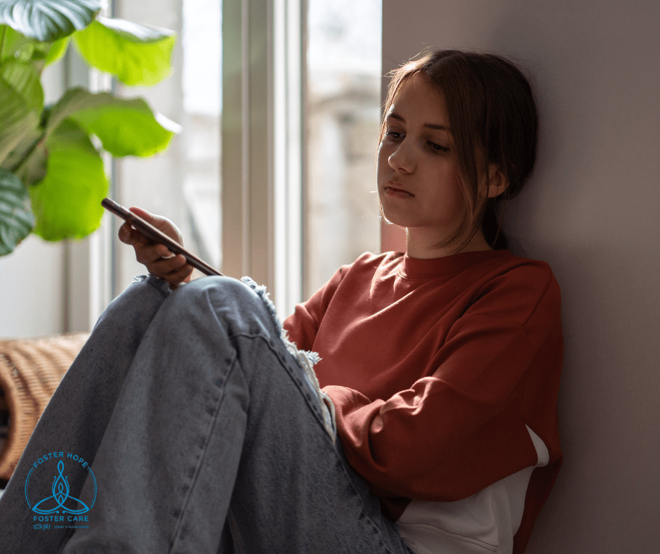 Teenager sitting by a window with a phone, looking thoughtful.