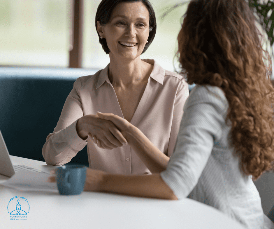 Two women shaking hands and smiling in an office setting, with a blue coffee mug and a laptop on the table.