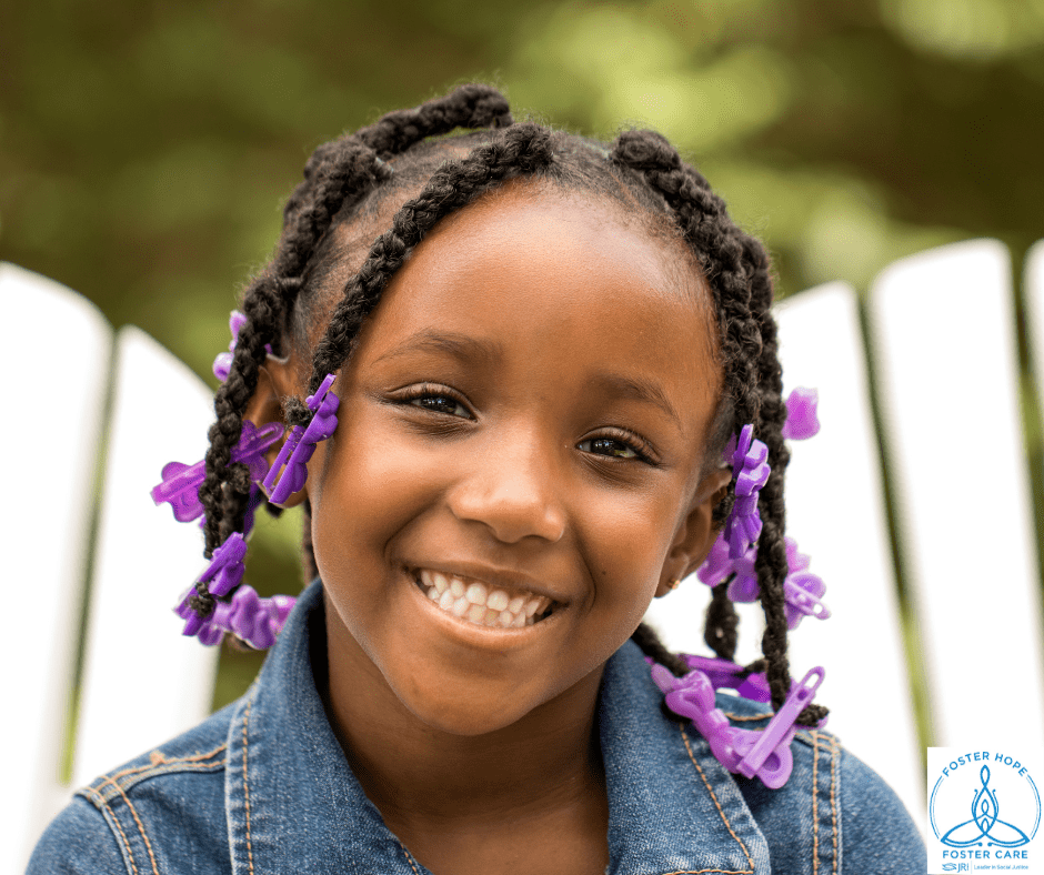 Girl with braided hair and purple clips smiling outdoors, wearing a denim jacket.