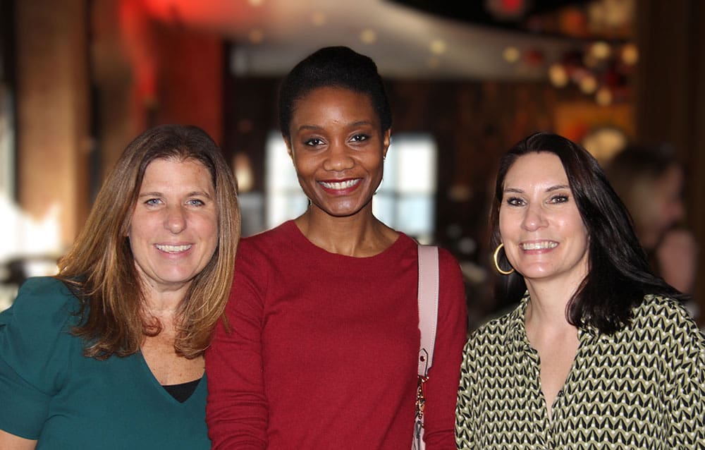 Three women smiling, standing close together indoors.