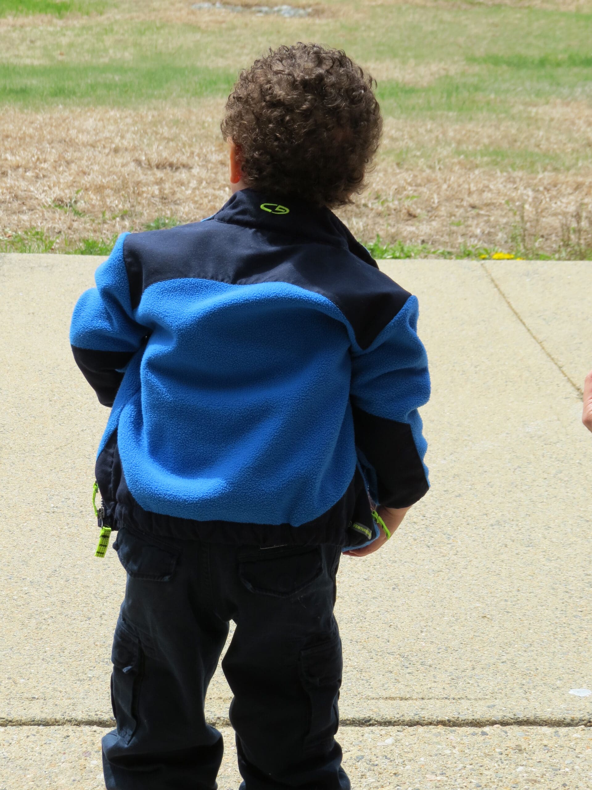 Child with curly hair wearing a blue and black jacket, standing on pavement.
