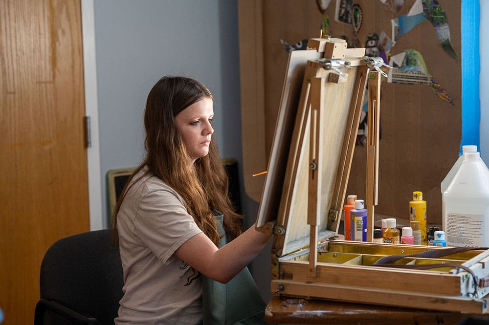 Person sitting at table, focused on painting on an easel.
