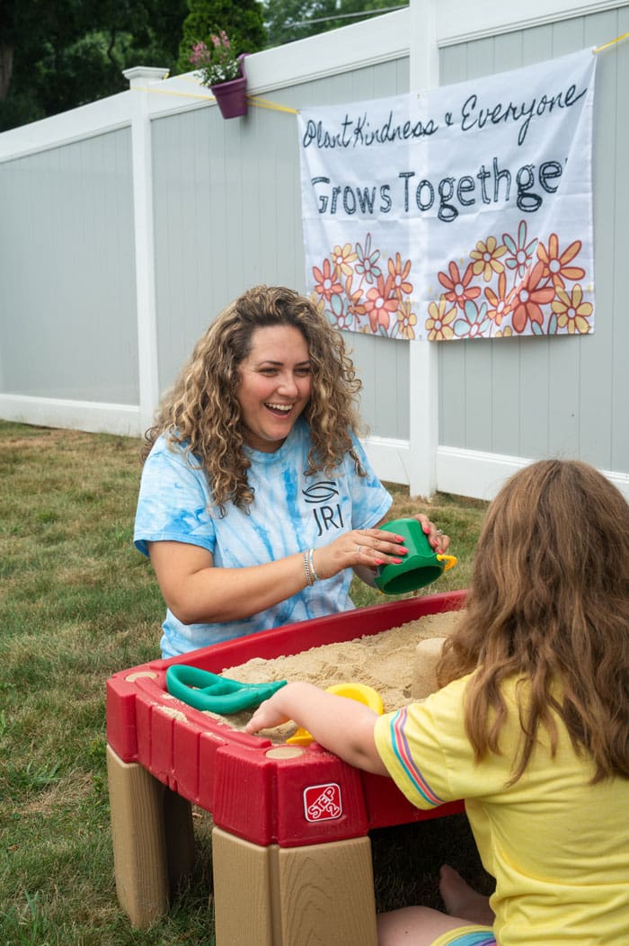 A person with curly hair smiling and playing in a sandbox with a child.