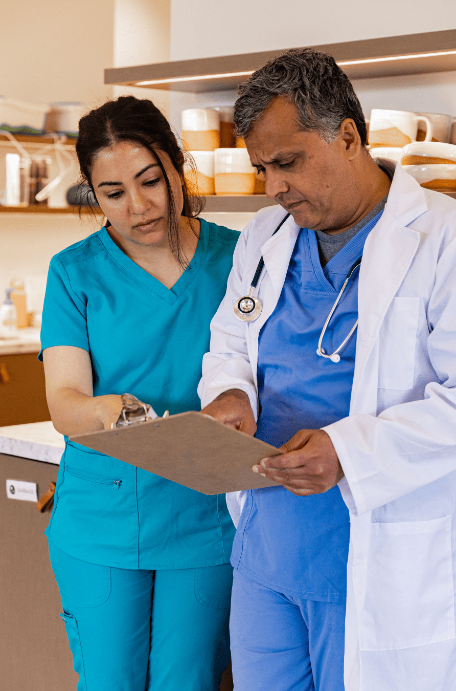 Two medical professionals, a woman in blue scrubs and a man in a white coat, review a clipboard together.