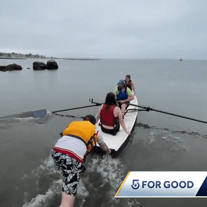 People in a boat with one person helping others to board in water with rocks nearby