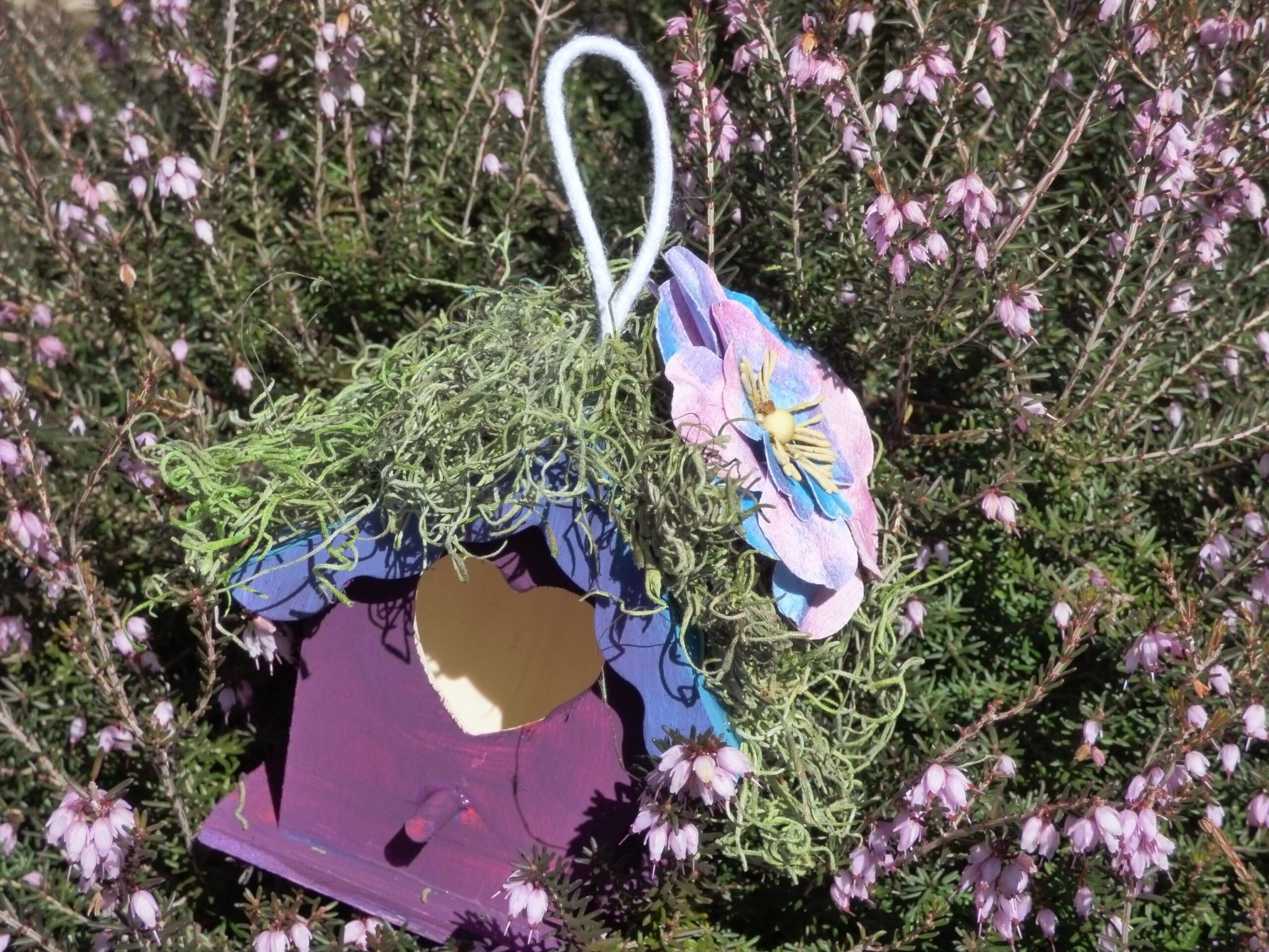Colorful decorative birdhouse with flowers hanging on it, surrounded by pink flowers