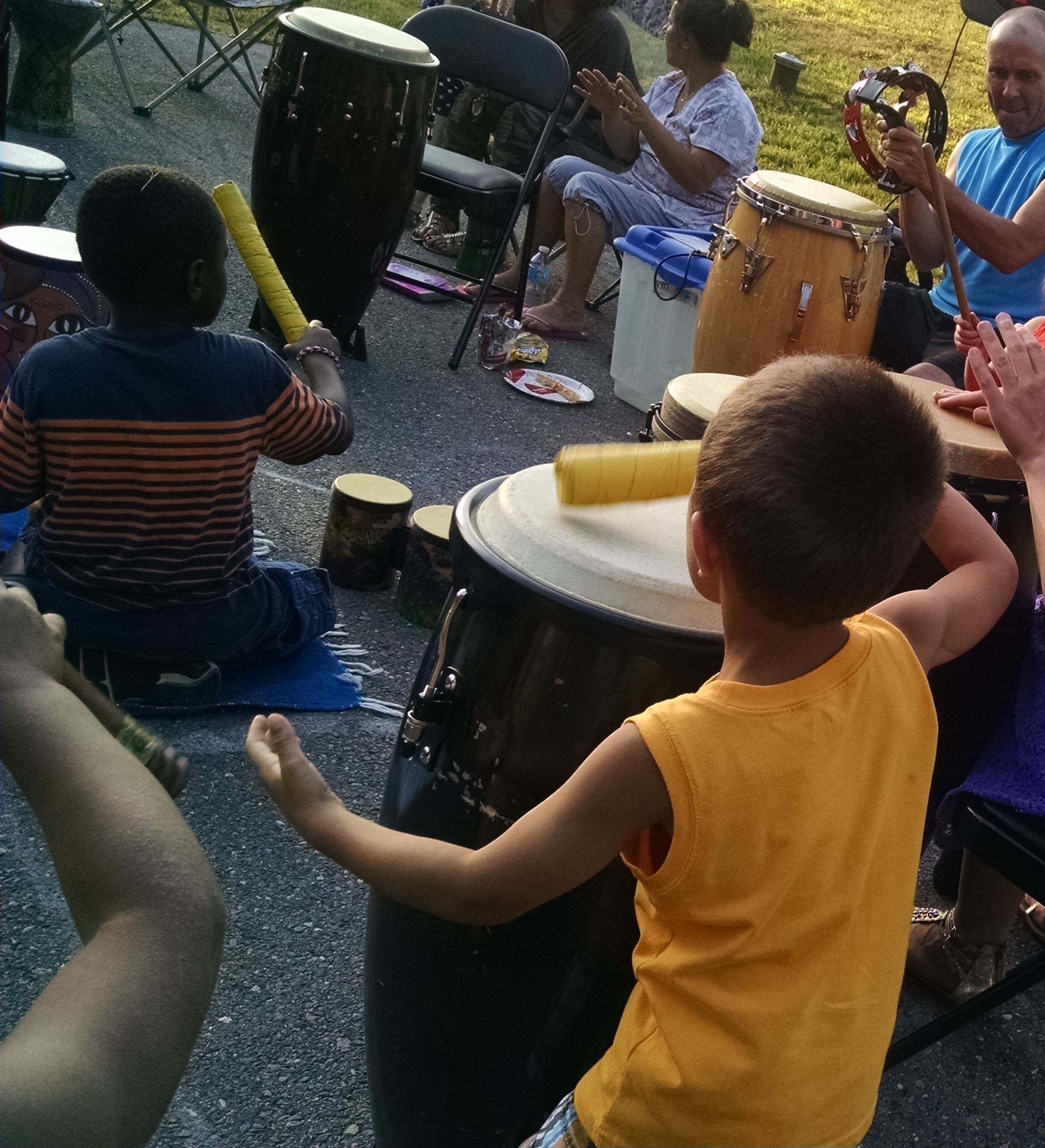Children playing drums during an outdoor event with adults in the background.