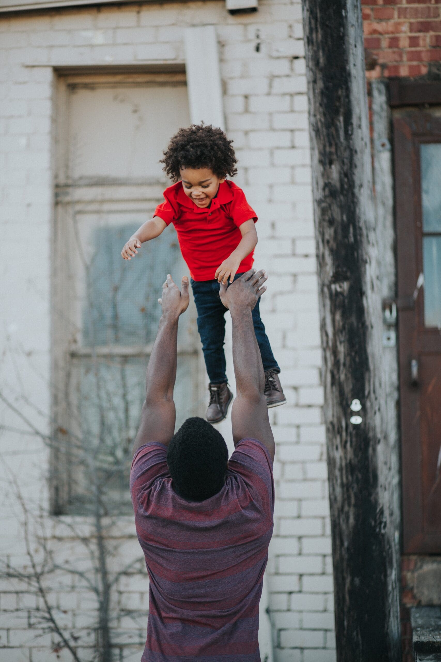 Parent lifting child with curly hair and red shirt outside.