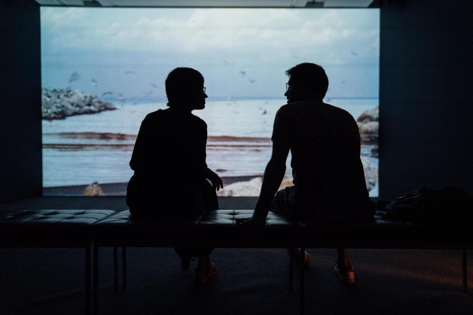 Two people sitting and talking in front of a large screen showing a beach scene.