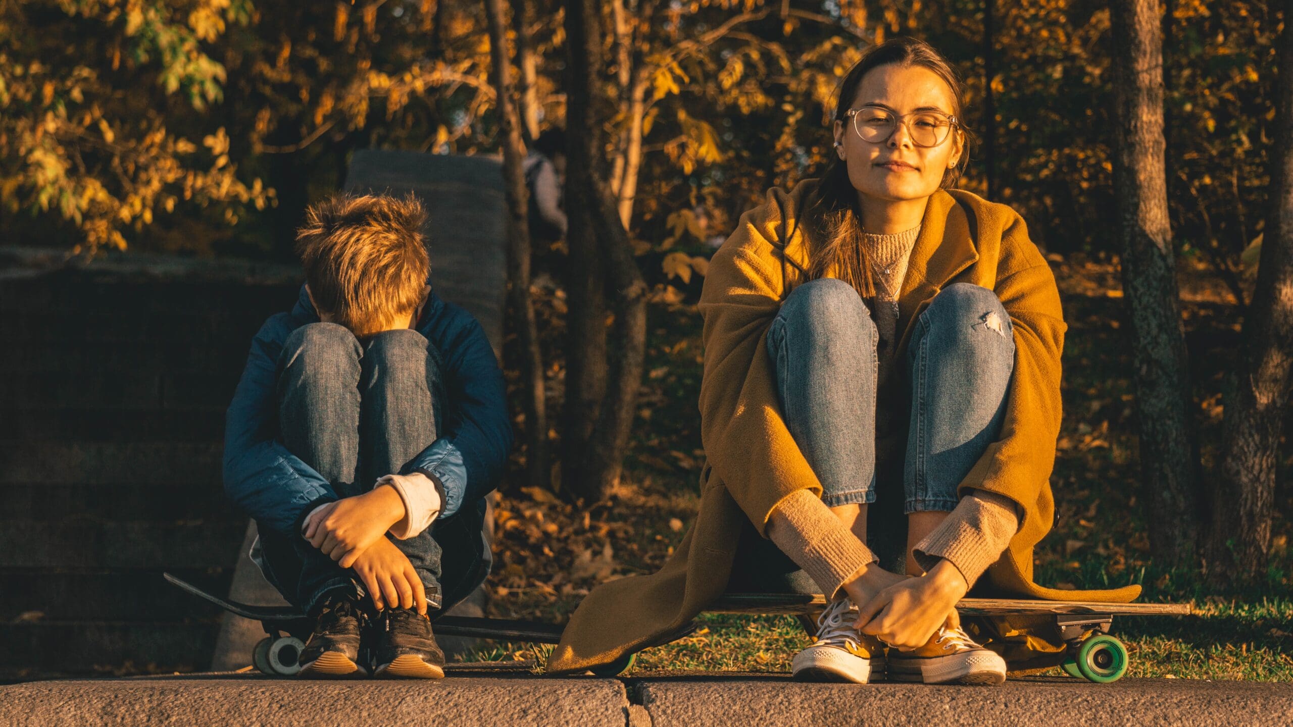 Adult and child sitting outdoors with skateboards, surrounded by trees.